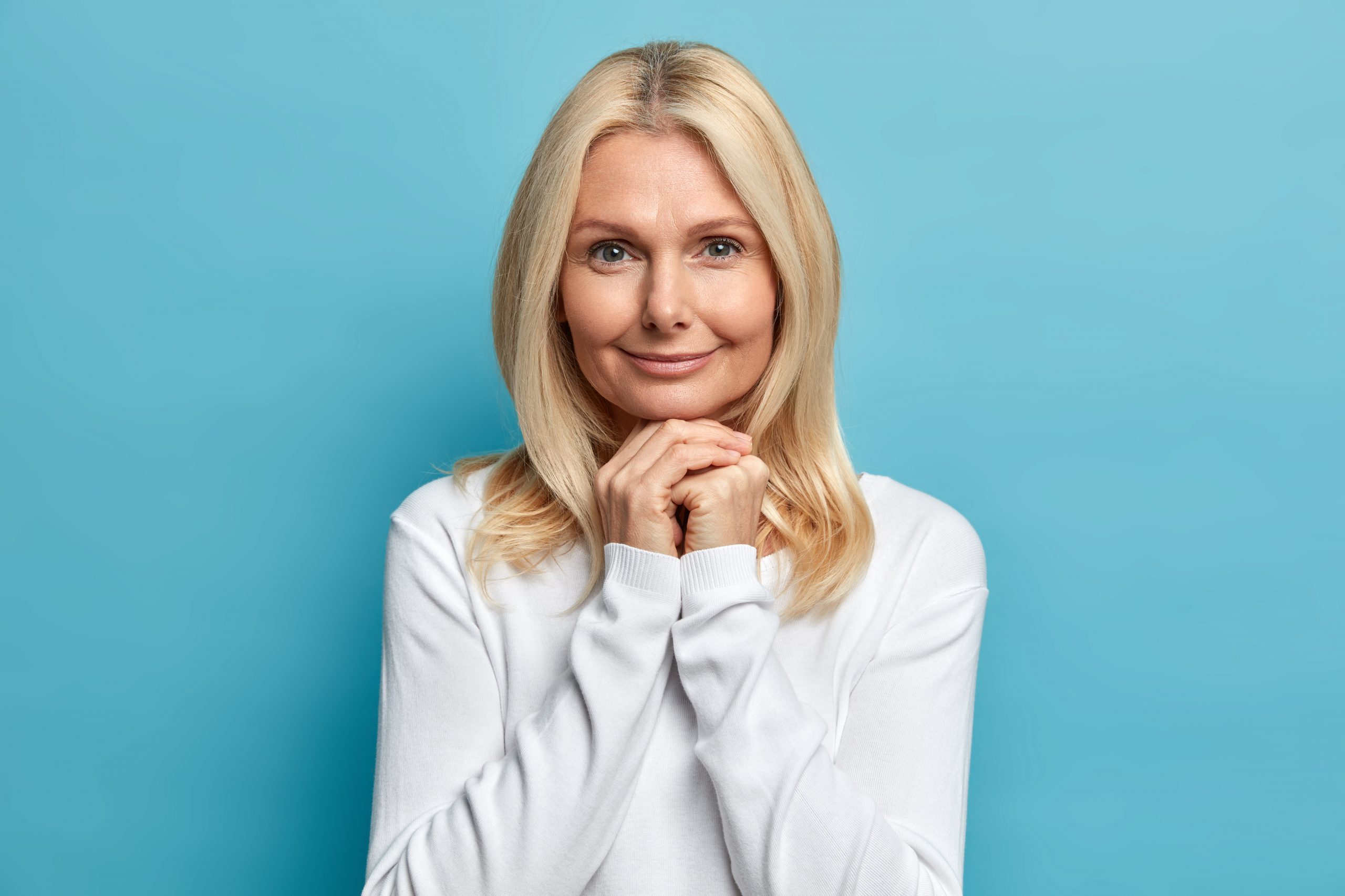 Studio portrait of confident fifty years old woman keeps hands under chin looks directly at camera with calm expression wears white sweater has well cared skin poses against blue background.